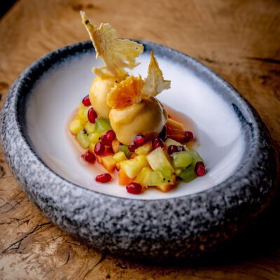 A plate of two scoops of mango sorbet, surrounded by a colorful mix of tropical fruits, including pineapple, kiwi, and pomegranate seeds