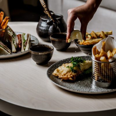A hand squeezes a lemon wedge over a plate of fish and chips, with a side of sandwiches and tea in the background.