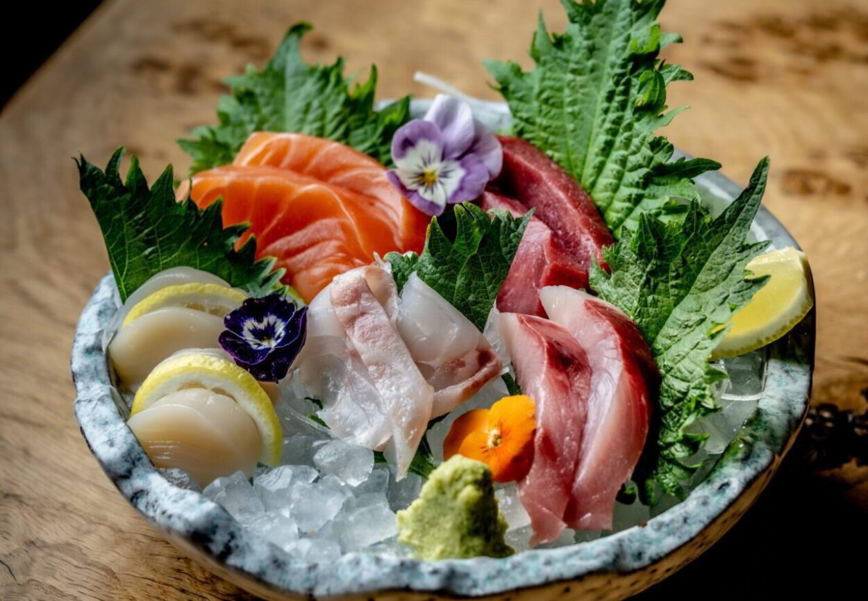 A plate of assorted sashimi, including salmon, tuna, and white fish, garnished with edible flowers, lemon wedges, and wasabi.