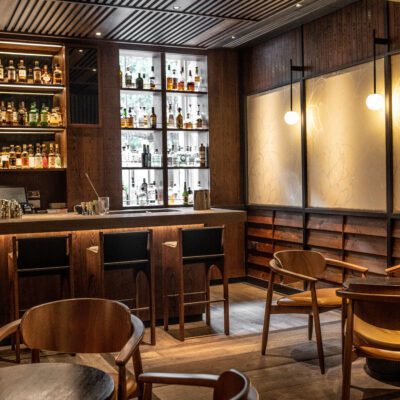 A dimly lit bar with a wooden counter, high chairs, and shelves stocked with liquor bottles.