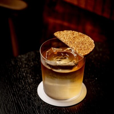 A cocktail served in a rocks glass with a large ice cube and a circular sesame cracker garnish resting on the rim