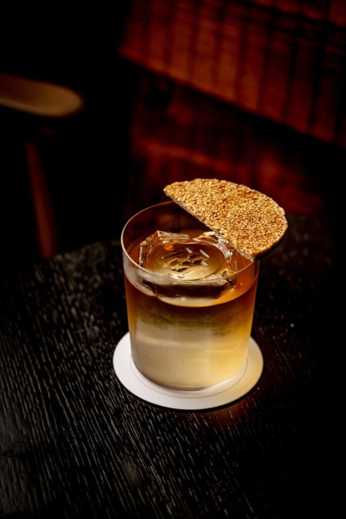 A cocktail served in a rocks glass with a large ice cube and a circular sesame cracker garnish resting on the rim