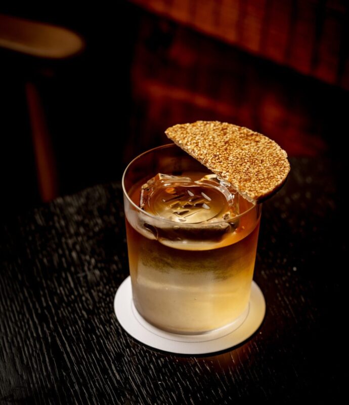 A cocktail served in a rocks glass with a large ice cube and a circular sesame cracker garnish resting on the rim