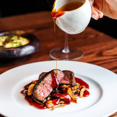 A plate of sliced steak with a rich sauce being poured over it. The steak is resting on a bed of sautéed mushrooms.